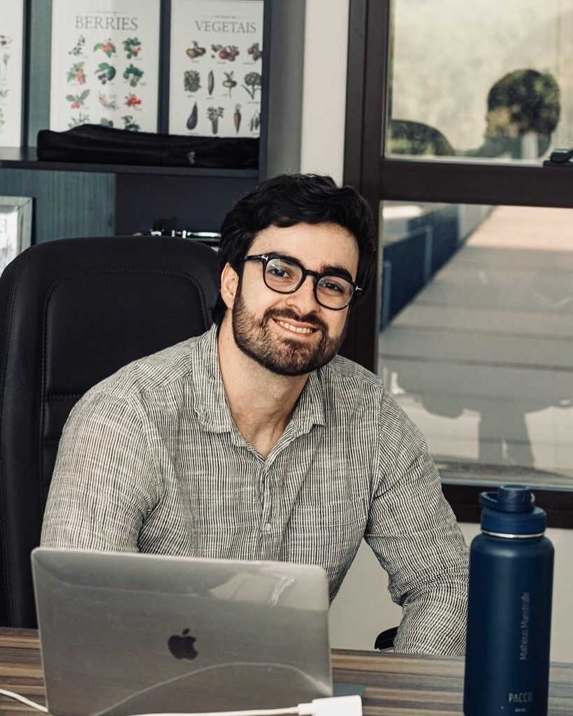Matheus Maestralli sorrindo, sentado à mesa do consultório, com o notebook e a garrafa de água